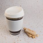 A reusable coffee cup in white with a silicone lid, alongside rolled wafer snacks on a speckled surface.