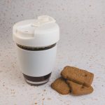 A reusable coffee cup in white, featuring a flip lid, alongside several biscuits on a speckled surface.