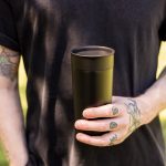 A stainless steel coffee cup in matte black, held by a person with tattoos, set against a natural background.
