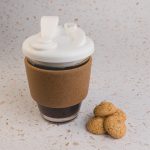 A reusable coffee cup with a cork band and white snap lid, beside three small cookies on a speckled surface.