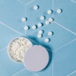 A mint tin containing small white mint tablets, with some tablets scattered around it on a light blue surface.