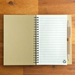 A recycled kraft notebook with lined pages, paired with a beige eco-friendly pen, featuring a logo.