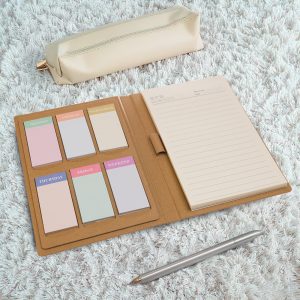 A planner with a beige cover, various coloured sticky notes, a notepad, and a silver pen on a textured surface.