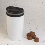 A reusable white coffee cup with a black lid, accompanied by several brown chocolate coffee drops.