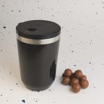 A reusable coffee cup in matte black with a stainless steel band and lid, accompanied by a few chocolate-covered treats.
