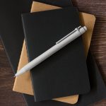 A grey pen rests on top of two notebooks, one black and one brown, on a wooden surface.