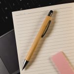 A bamboo pen with a black stylus tip, placed on a lined notepad next to a pink sticky note and a laptop.