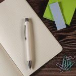 A white pen rests on a dotted notebook, accompanied by colourful sticky notes and paperclips on a wooden surface.