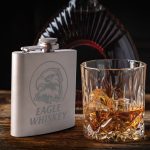 A stainless steel hip flask with an engraved design beside a glass of whiskey containing ice.