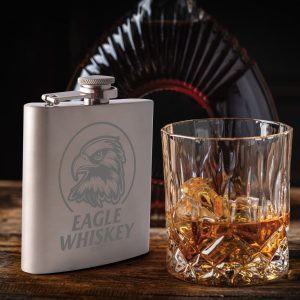 A stainless steel hip flask with an engraved design beside a glass of whiskey containing ice.