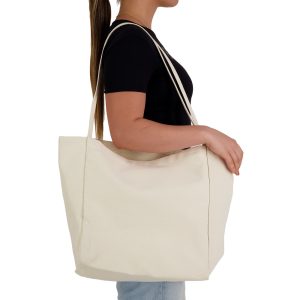 A beige tote bag with long handles, carried by a person, features a minimalist design and a logo.