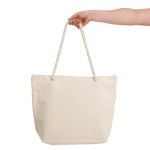 A hand holds a large, cream-coloured canvas tote bag with rope handles. The bag is plain and unbranded.