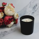 A medium black glass candle with a lit wick, beside a bouquet of red and white flowers on a marble surface.