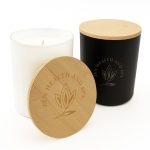 A large glass candle in black with a wooden lid, featuring an engraved logo design. The candle itself is white.