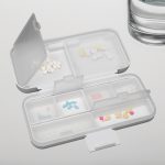 A compact, multi-section pill organiser in white, containing various coloured tablets, next to a glass of water.