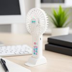 A white handheld fan with a base, featuring a logo and colourful text, placed on a desk near greenery and office supplies.