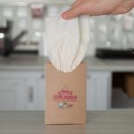 A hand holds a white tissue being pulled from a brown cardboard box with a playful logo.
