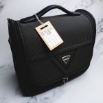 A black luggage bag with a wooden luggage tag attached. The tag has a logo and is designed for identification.