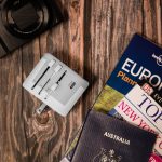 A white travel adaptor positioned on a wooden surface, surrounded by a camera, travel books, and a passport.