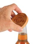 A drink bottle opener coaster in bamboo with a hand positioned above it, showing its natural wood color and branding.