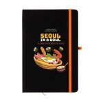 A black A5 notebook with an illustrated cover featuring food items and a logo. It has an orange elastic band.