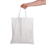 A white cotton tote bag with short handles is held by a hand. The bag features a simple design and a blank surface.