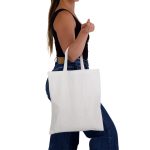 A woman carries a plain white cotton tote bag with short handles, wearing casual jeans and a black top.