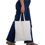 A white cotton tote bag with short handles, being held by a person wearing blue jeans.