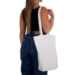 A white cotton long handle bag is slung over the shoulder of a person wearing a black top and denim jeans.