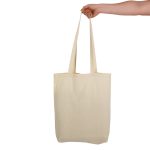 A cream-coloured tote bag with long handles held by a hand. The bag includes a logo.