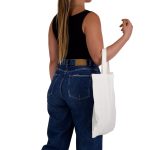 A woman with long hair holds a plain white short handle bamboo tote bag while wearing a black top and blue jeans.