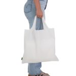 A white reusable grocery bag is being held by a person wearing blue jeans and shoes. The bag features a logo.