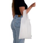 A white reusable grocery bag is held by a person wearing a black top and light blue jeans, with a visible logo.