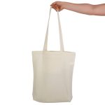 A cream-coloured tote bag with long handles and a pocket, held by a hand. It features a logo.