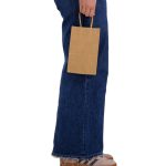 A small brown paper bag held by a person wearing blue jeans and light-coloured shoes. The bag has handles and an embossed logo.