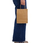 A medium brown paper bag with handles is held by a person wearing jeans. The bag has a plain design.