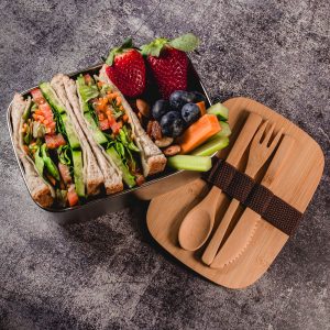 A stainless-steel lunch box containing wraps, strawberries, blueberries, celery, and bamboo cutlery with a strap.