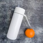 A drink bottle in white stainless steel with a clear straw attachment, placed next to an orange.