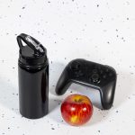A drink bottle featuring a flip straw lid, alongside a red apple and a black gaming controller.