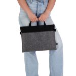A grey and black felt satchel is held by a person wearing light blue jeans, featuring top handles for easy carrying.