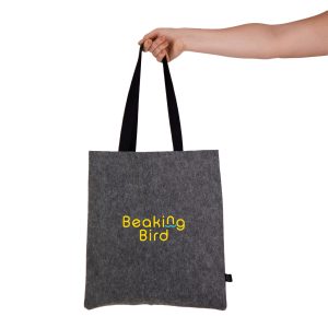 A grey felt tote bag with black straps is held by a hand, featuring a logo design.