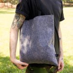 A grey felt tote bag held by a person in a black shirt, featuring a strap and a logo.