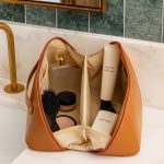 A tan toiletry bag with compartments containing a brush, hand cream, lotion, and jars of products, featuring a logo.