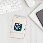 A beige notepad with a spiral binding and a pen, featuring a logo on the front, on a white desk.