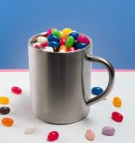 A stainless steel mug filled with assorted colour mini jelly beans, with some jelly beans scattered around the base.