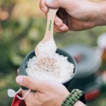 A hand holds a beige spoon scooping rice from a gray bowl outdoors. The spoon features a logo.
