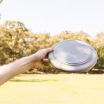 A hand holds a gray frisbee against a sunny outdoor backdrop of trees and grass.