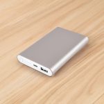 A silver power bank with a USB port on a wooden surface, featuring a sleek, minimal design and a logo.