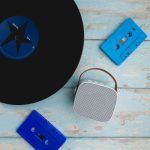A Boombox Mini Speaker is placed on a rustic surface alongside a vinyl record and two blue cassette tapes.