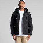 A man wearing a black canvas heavy hooded jacket with zip and front pockets. The jacket has a hood and a grey lining.